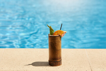 Exotic cocktail in wooden glass with lemon and drinking tube standing on poolside with blue pool's...