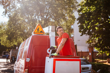  A caucasian worker in a red T-shirt stands in a basket in a crane . Setting up traffic lights in progress