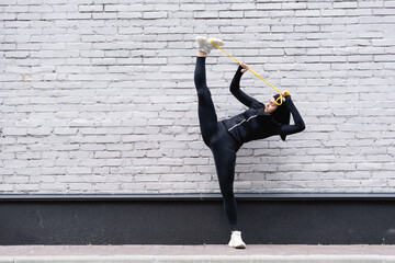 flexible arabian sportswoman in hijab exercising with resistance band near brick wall