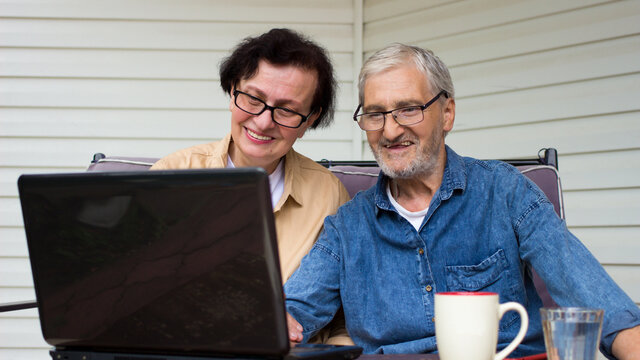 Portrait of smiling elderly couple using laptop computer,sitting on home terrace veranda.Happy senior caucasian mature and retired man and woman using technology,wireless internet connection at home