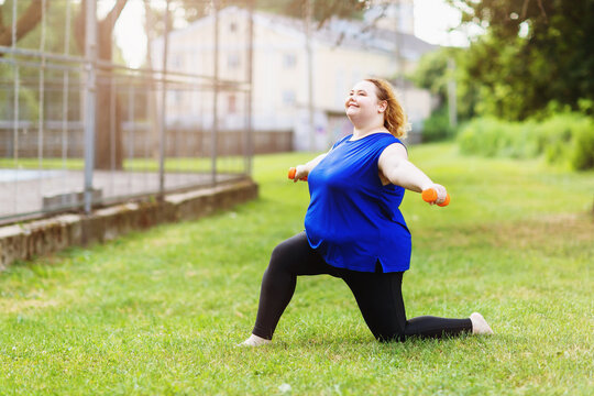 Young positive blonde size plus performs exercises with dumbbells in the park. The concept of a healthy lifestyle