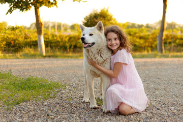 Frontal portrait of a little cute girl with a big white dog in the park. A beautiful 7 year old girl in pink dress hugs her favorite dog during a summer walk.