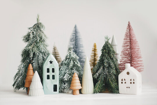 Christmas little houses and trees on white background. Festive modern decor. Happy holidays. Miniature cozy village, ceramic houses, wooden and handmade christmas trees. Space for text