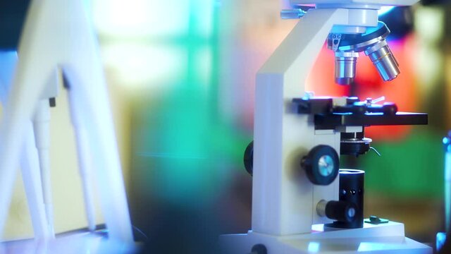 Camera tracks right past a line of out of focus laboratory paraphernalia in the foreground to an in focus microscope in the background.