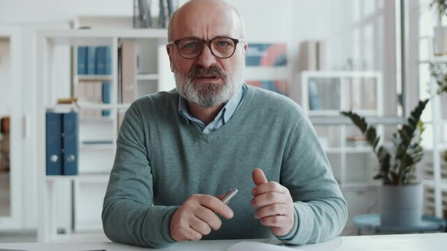 Senior businessman in wireless earphones sitting at desk in the office, looking at camera and speaking while discussing job via online call