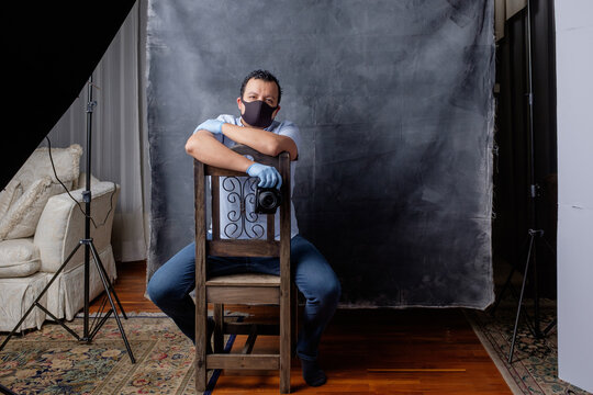 Environmental photo of a young portrait photographer with a retro camera sitting down in his studio. He is wearing a face mask and surgical gloves.