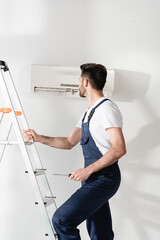 workman holding screwdriver while standing on stepladder near air conditioner