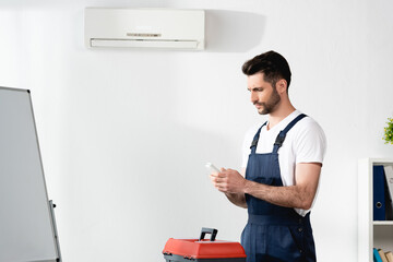thoughtful repairman holding remote controller while standing near toolbox and air conditioner