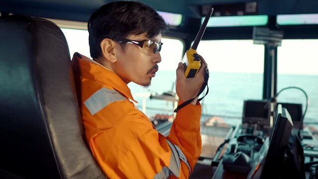 Filipino deck Officer on bridge of vessel or ship wearing coverall during navigaton watch at sea . He is speaking on GMDSS VHF radio, communication between vessels.