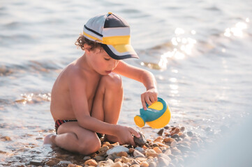 Little boy on a pebble beach. a little boy sits on the beach and plays with a watering can