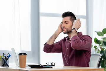dreamy businessman with closed eyes touching wireless headphones while sitting at workplace near coffee to go