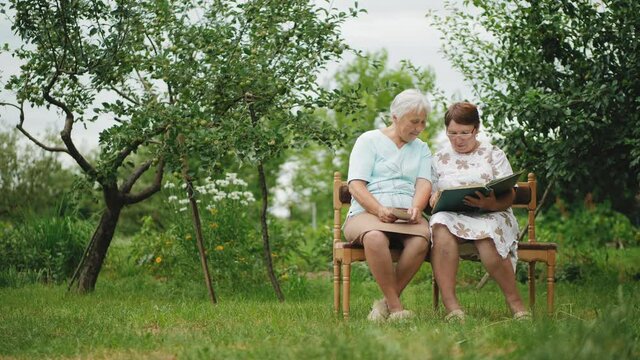 Two older women view photos in an old family album sitting on a bench in a green garden.