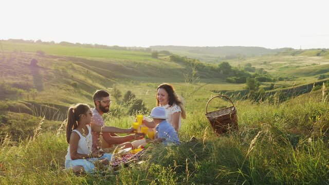 A family of four have fun together on a picnic in nature, children and parents smile, eat and drink