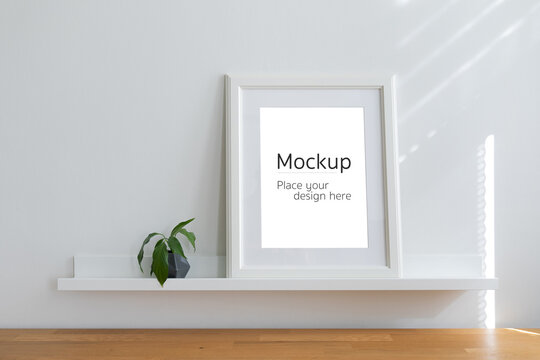Vertical white mockup frame standing on small shelf with potted plant. Cardboard passepartout with free space inside of it. Sun beams on the wall behind the workplace.