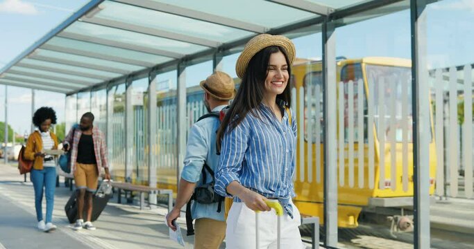 Caucasian young beautiful woman in hat walking outdoor and carrying suitcase on wheels. Pretty female tourist strolling at bus and tram stop and smiling. saying hello or hi to handsome guy.