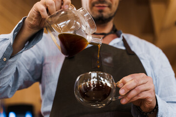 Closeup pouring coffee in double glass cup in cafe by handsome bearded barista. Coffee brewing syphon and aeropress alternative methods. Advert for social networks for cafe and restaurant.