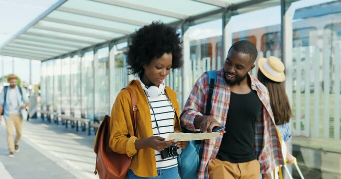 African American young couple of tourists walking outdoors with map and looking for right route. Beautiful woman and handsome man travellers talking and discussing way to choose with city plan.