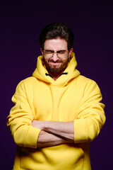 A young man of 25-30 years in glasses and a yellow sweatshirt emotionally poses on a purple background. 