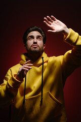 A young man of 25-30 years old in a yellow sweatshirt emotionally pulls his hands up on red wall background. 