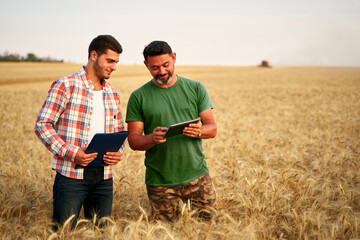 Naklejka na ściany i meble Two farmers stand in wheat stubble field, discuss harvest, crops. Senior agronomist with touch tablet pc teaches young coworker. Innovative tech. Precision farming with online data management soft.