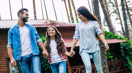 Young happy modern parents with a cute daughter are walking outdoors and holding hands together on suburban house background.