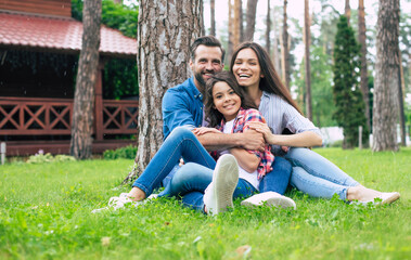 Naklejka na ściany i meble Beautiful happy family while sitting together on the grass and hugging each other, relaxing outdoors on suburban house background.
