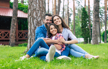 Naklejka na ściany i meble Beautiful happy family while sitting together on the grass and hugging each other, relaxing outdoors on suburban house background.