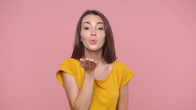 Smiling cute young woman girl 20s in yellow t-shirt posing isolated on pink background studio. People lifestyle concept. Look camera charming smile send blow air kiss hands heart sign say i love you