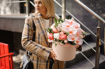 young blonde woman holds round pink box with floral arrangement of peonies and orchids and looks away