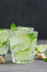 Fresh summer mojito drink with lime slices, mint, ice cubes and brown sugar in a glass on a dark background. Vertical, copy space.