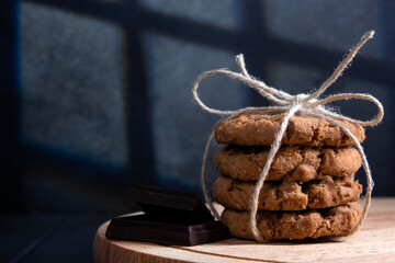Fototapeta na wymiar Oatmeal cookies tied with coarse rope with chocolate slices on wooden board. Dark background with incident light from the window frame.