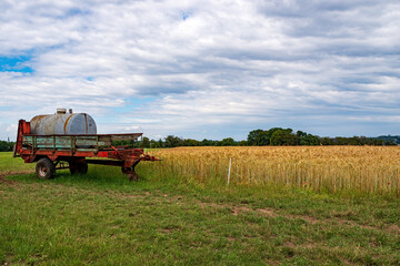 View over a barley field with dramatically cloudy sky, on the left an old water tank on a trailer