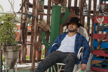 Obraz na płótnie Canvas Hombre joven con camisa a cuadros y sombrero leyendo sobre la terraza, y usando binoculares 