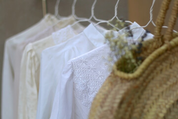 Clothing rack with white and neutral garments and wicker bag with flowers. Selective focus, gray concrete background.
