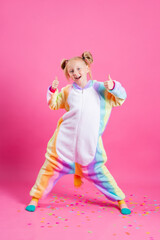 Happy little girl in kigurumi unicorn on a pink background rejoices in multi-colored confetti, space for text