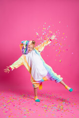 Happy little girl in kigurumi unicorn on a pink background rejoices in multi-colored confetti, space for text