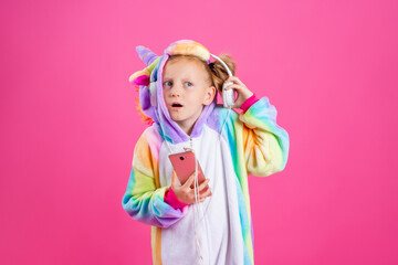 Fototapeta na wymiar Happy little blonde girl in unicorn kigurumi listens to music holding in hand smatrphone on a pink background