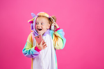 Happy little blonde girl in unicorn kigurumi listens to music holding in hand smatrphone on a pink background
