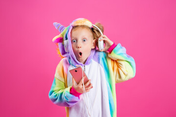 Obraz na płótnie Canvas Happy little blonde girl in unicorn kigurumi listens to music holding in hand smatrphone on a pink background