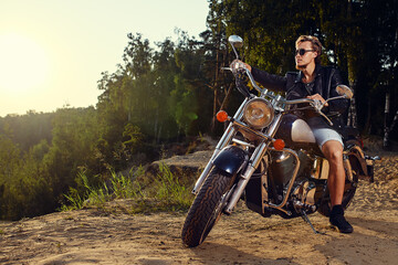 Fototapeta premium Brutal young man in sunglasses, blue jeans and a black leather jacket sitting on the custom motorcycle