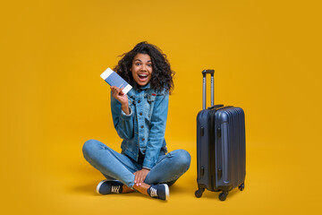 Full-length portrait of smiling tourist girl sitting with her suitcase, holding passport and boarding pass and looking directly to the camera with happy face expression
