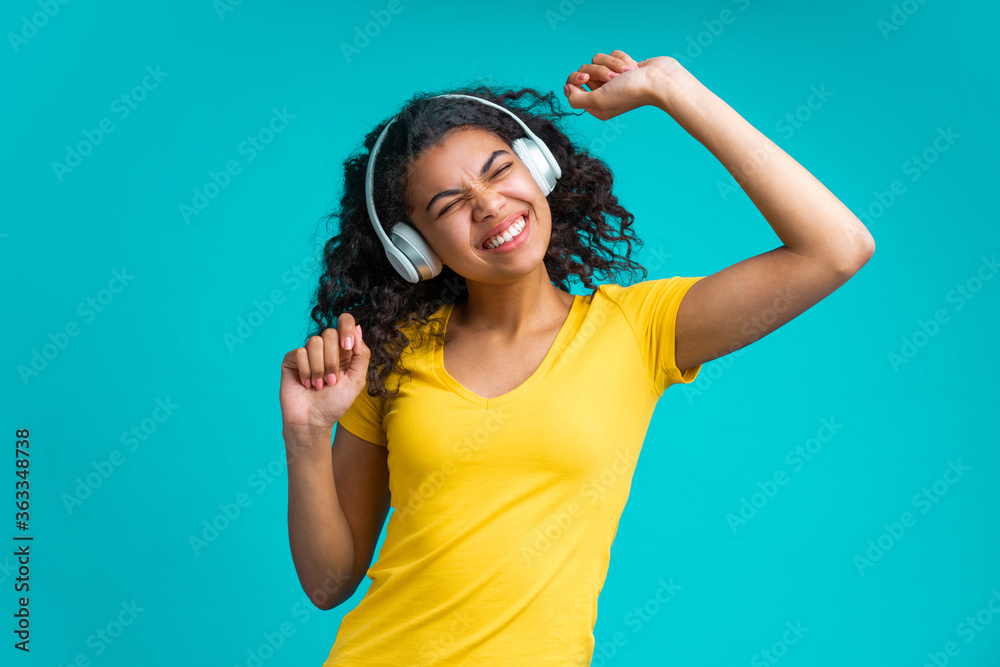 Wall mural Cute dark skinned girl having fun enjoying her day-off singing and dancing while listening music via wireless headset over blue background