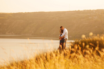 Romantic couple, man embracing woman while standing on a field near a lake at sunset