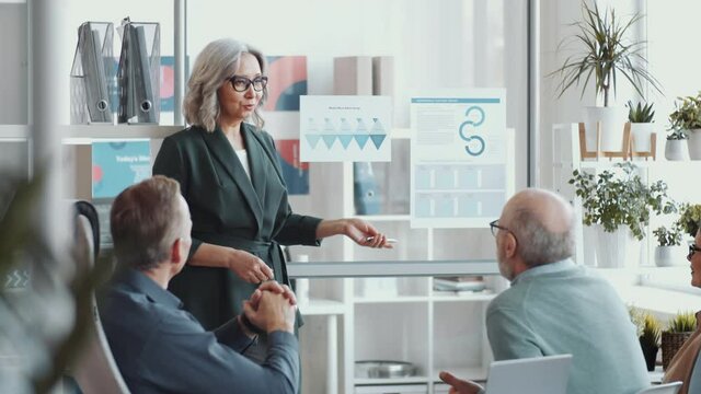 Senior female business trainer pointing at posters on glass wall, smiling and giving a speech in front of mixed-aged workers during educational seminar in the office