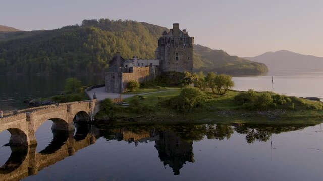 Aerial drone shot tracking around the stunning Eilean Donan Castle in Scotland at sunset, with the lake surrounding it and mountains in the background, and the reflection in the water.