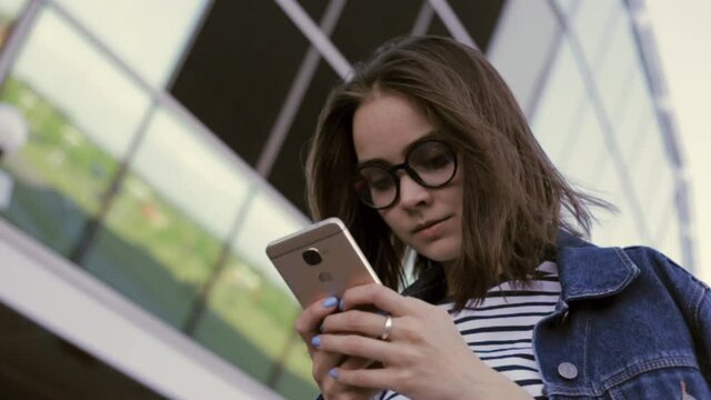 Low angle of young woman in eyeglasses chatting on social media and writing messages on smartphone while standing on street near modern glass building