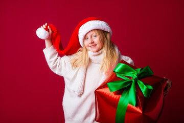 Happy little blonde girl in a santa hat holding a New Year's gift on a red background. New Year's sales. Space for text