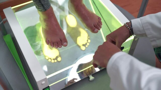 Close-up shot of special orthopedic equipment for feet examination. Female patient staying barefeet on transparent glass surface with green neon light while orthopedist physician examining her foot