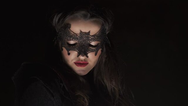 Beautiful queen in cloak with eyes mask at dark background. Halloween. Portrait of young brunette vampiress. Close-up in 4K, UHD