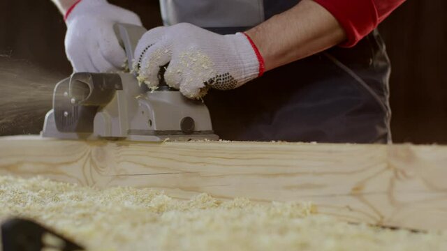Close-up hands of joiner using electric planer with wooden blank in workshop. Work with electric planer for polish boards and bar. Wooden furniture manufacturing concept.